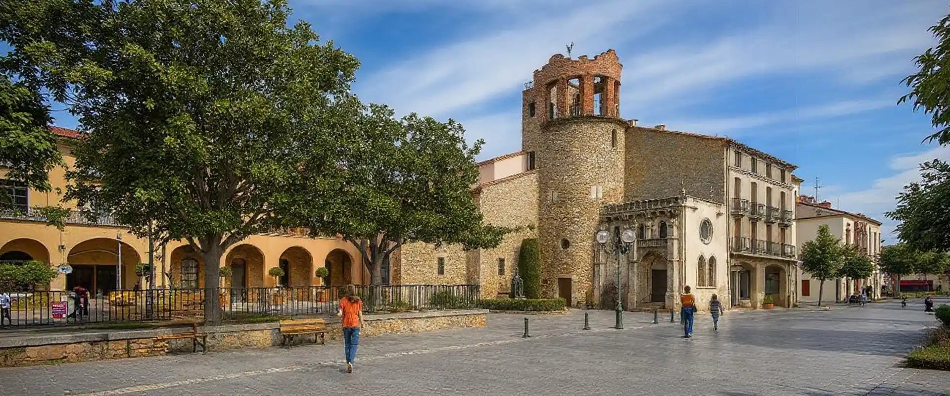 Une place pavée avec quelques promeneurs, bordée d'arbres et de bâtiments historiques - comme la tour en pierre avec des arcs et une horloge - reflète le charme de Thuir, où l'artisanat de Couvreur Thuir est visible sous un ciel bleu avec de légers nuages.