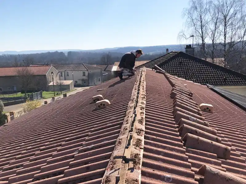 Une personne est accroupie au sommet d&#039;un toit de tuiles rouges, occupée à des travaux de couverture, avec des maisons et des arbres visibles à l&#039;arrière-plan sous un ciel clair.